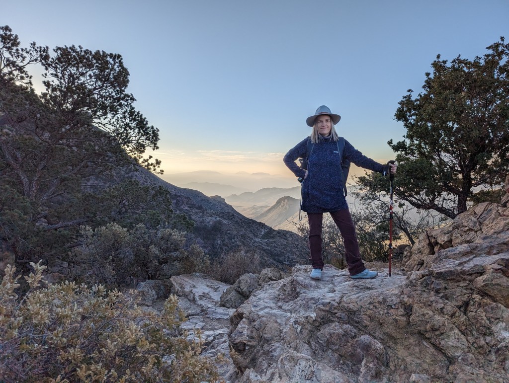 Petite woman dressed in winter gear with trekking poles standing atop a rocky trail with mountains visible in the background. 